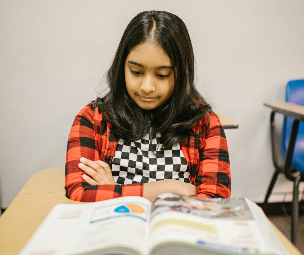 A focused young girl reading a book at her desk in a classroom setting.